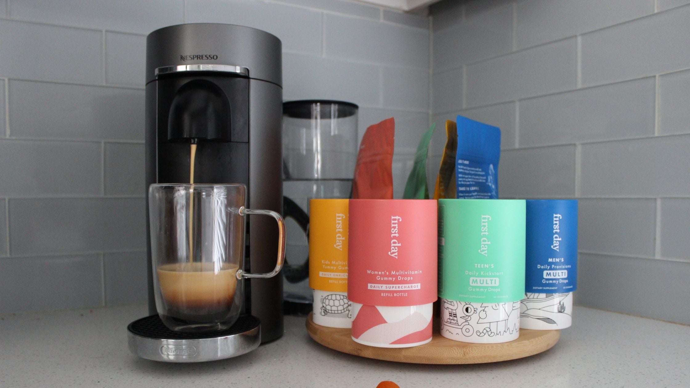 Kitchen countertop scene with a Nespresso machine brewing coffee and colorful 'first day' multivitamin bottles on a wooden tray