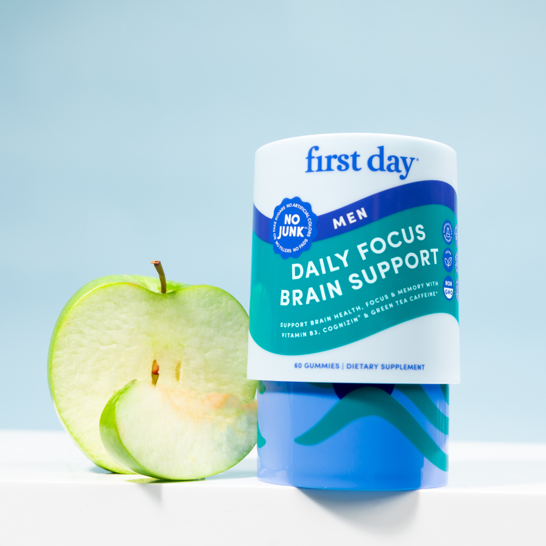 A container labeled First Day Men Daily Focus Brain Support stands beside a sliced green apple. The container is blue and white with wellness claims, set against a light blue background.