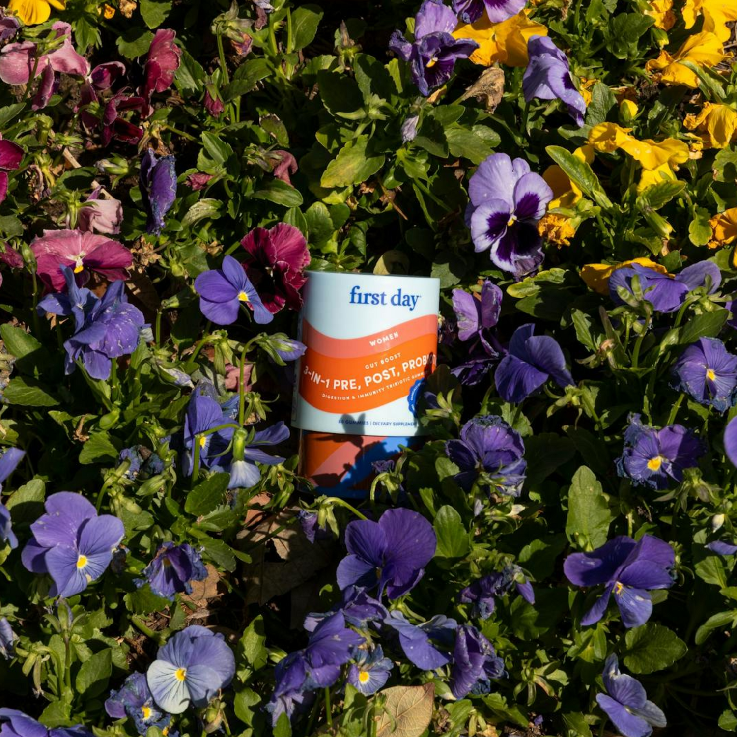 A container of First Day Women’s 3-in-1 Pre, Post, Probiotics sits nestled among vibrant purple, yellow, and maroon pansy flowers and green leaves.