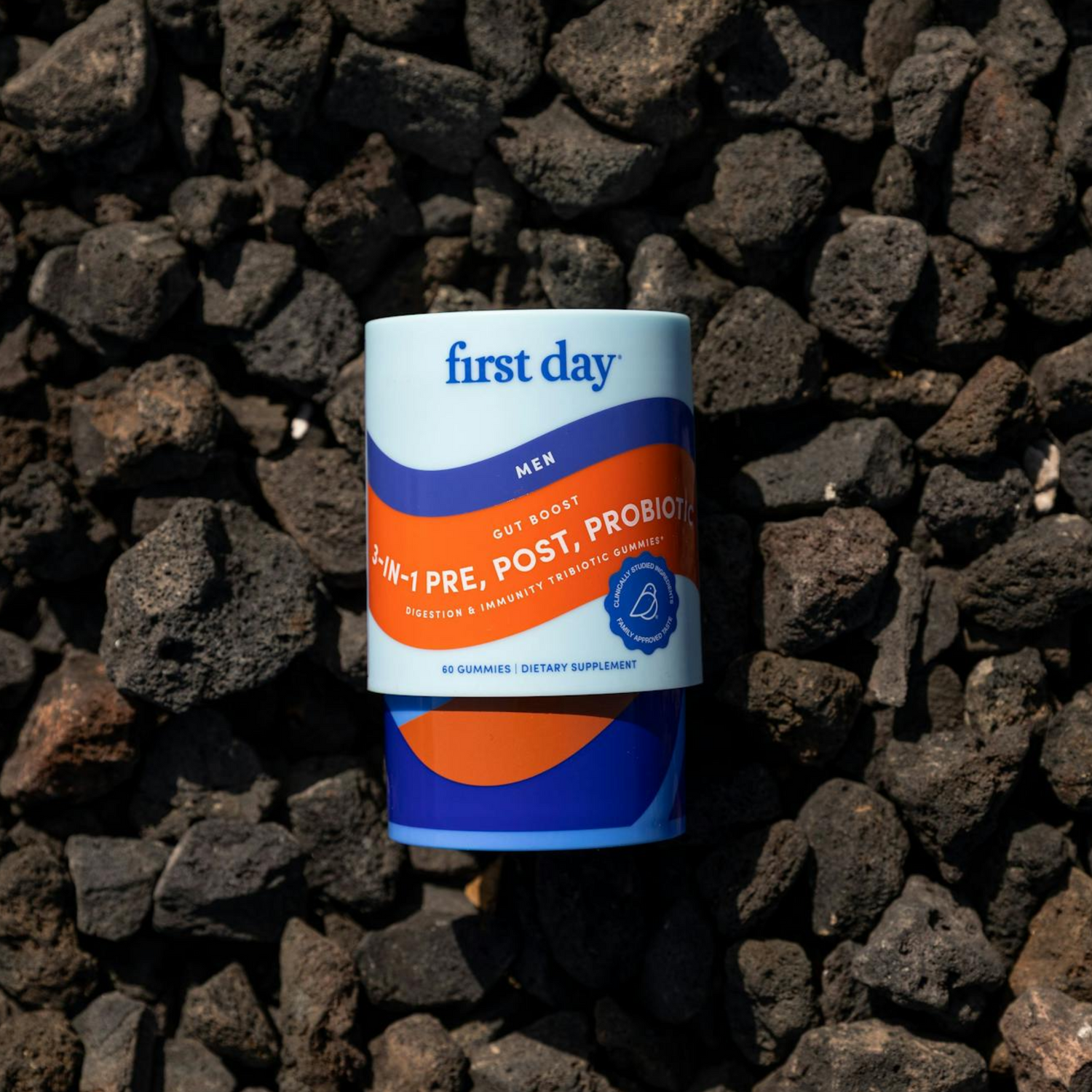 A container of first day 4-in-1 Pre, Post, Probiotics for men rests on a surface of dark, uneven volcanic rocks. The packaging is blue and orange with white and orange text.