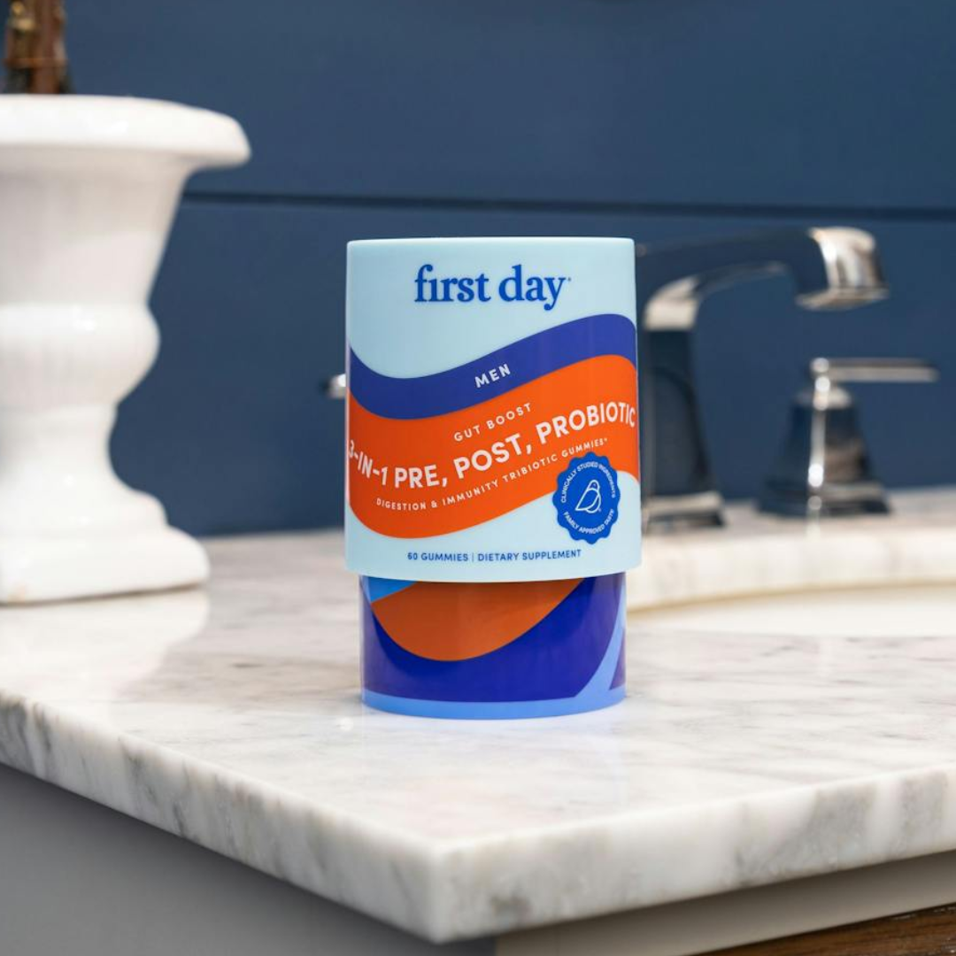 A container of First Day Mens 3-in-1 Pre, Post, Probiotic gummies sits on a marble bathroom countertop near a sink with a blue wall in the background.