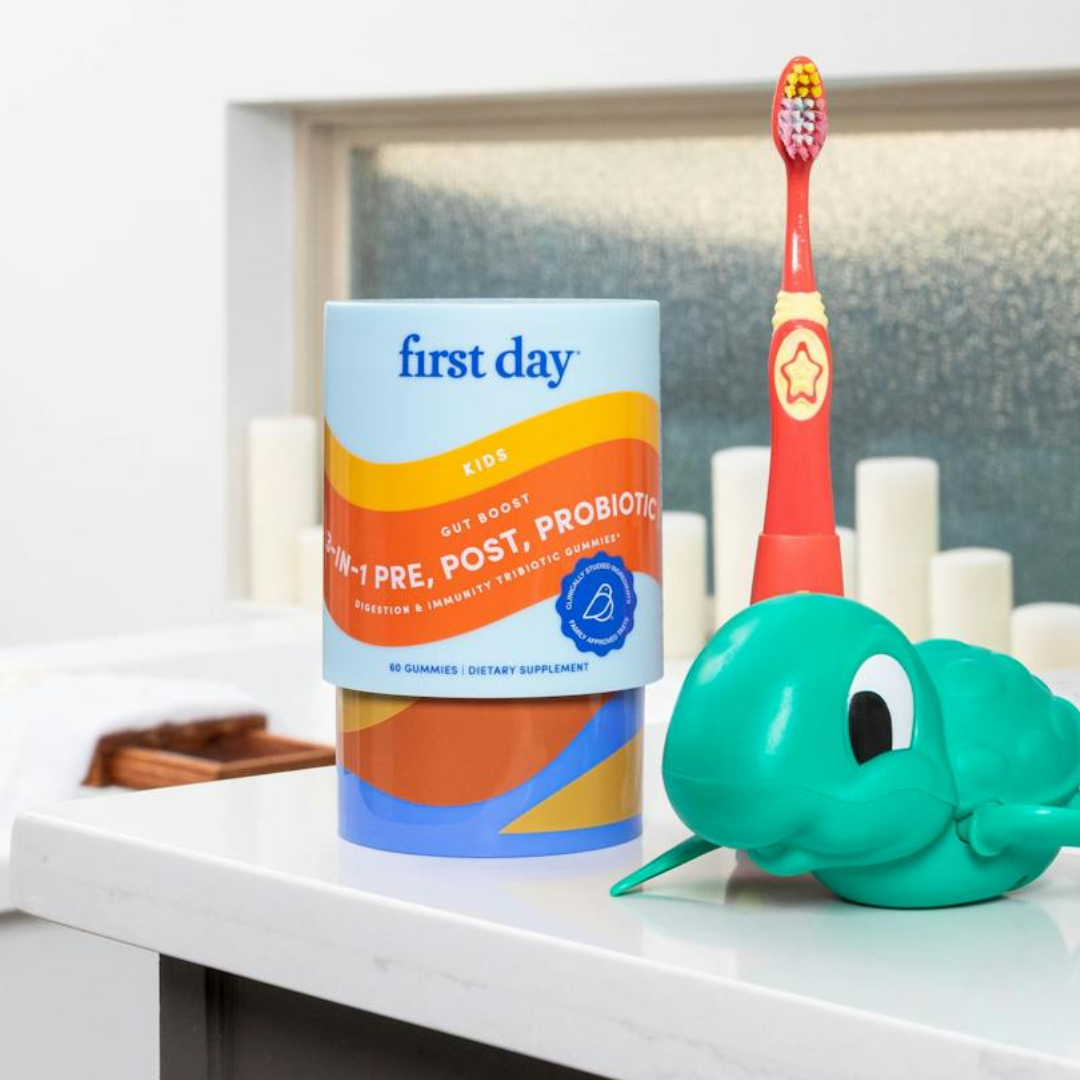 A container of First Day Kids 3-in-1 Pre, Post, Probiotic gummies sits on a bathroom counter next to a red childrens toothbrush and a green turtle-shaped toy. White candles are in the background.