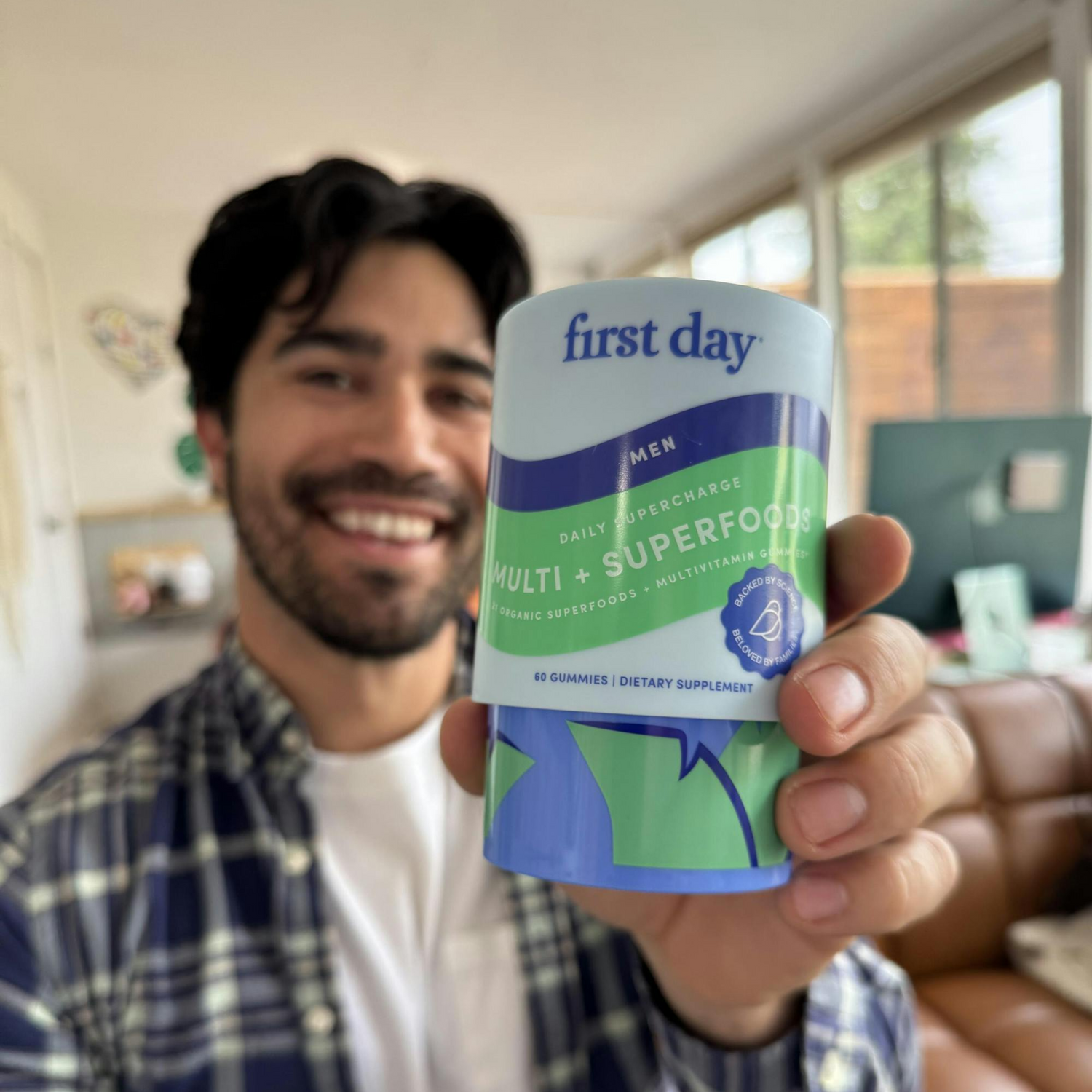 A smiling man holds up a container of first day Mens Multi + Superfoods gummies, with the product label clearly visible in a bright, modern living room.