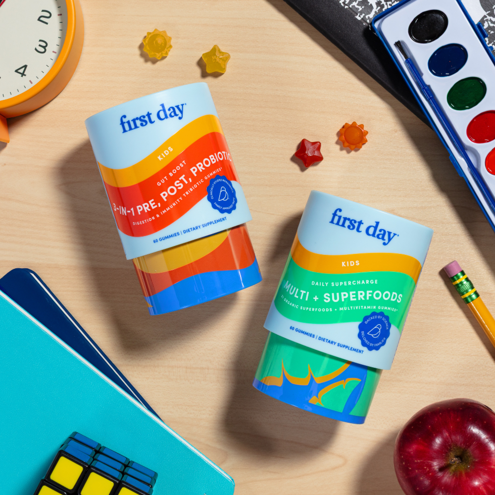 Two colorful First Day kids supplement bottles sit on a wooden desk surrounded by a Rubik’s cube, a red apple, a blue notebook, a pencil, watercolor paints, and star-shaped erasers.