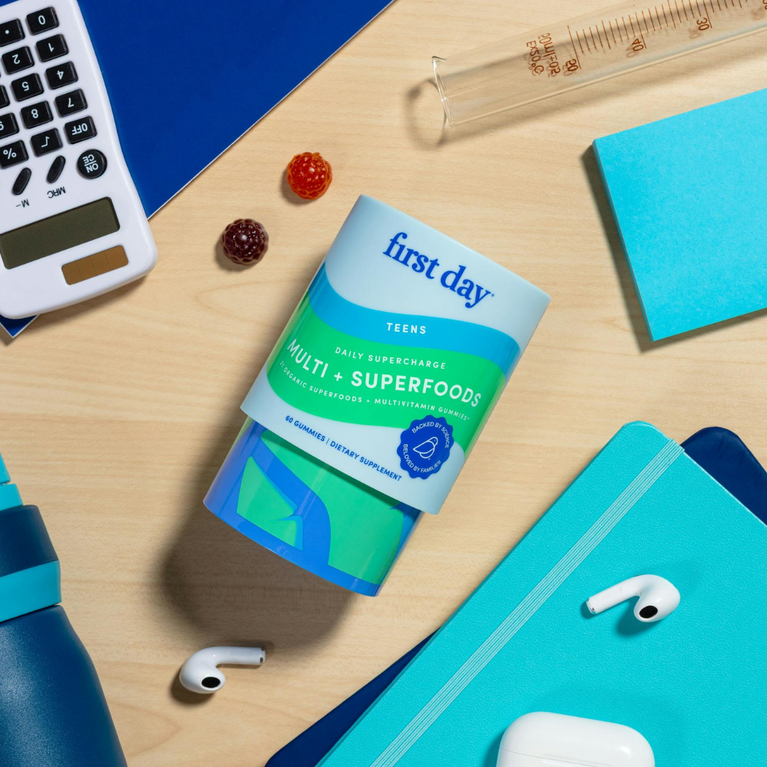 A container of First Day Teens Multi + Superfoods gummy vitamins lies on a desk surrounded by a calculator, notepad, water bottle, AirPods, sticky notes, and two gummies.