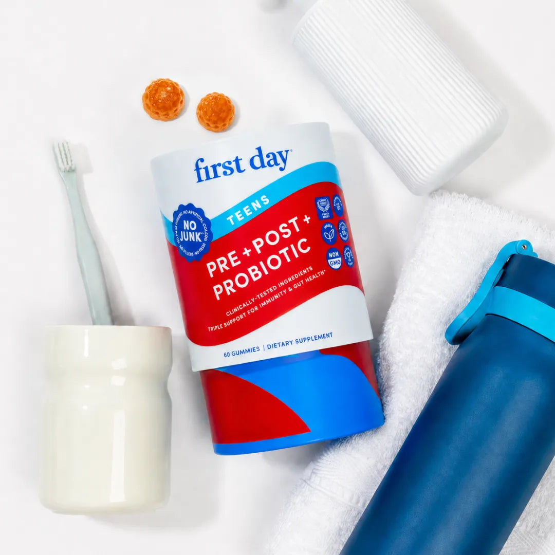 A First Day Tees Pre & Post & Probiotic supplement bottle is displayed in a flat-lay arrangement with dental and hydration props, including a toothbrush, toothpaste tube, toothpick holder, and water bottle, on a white background.
