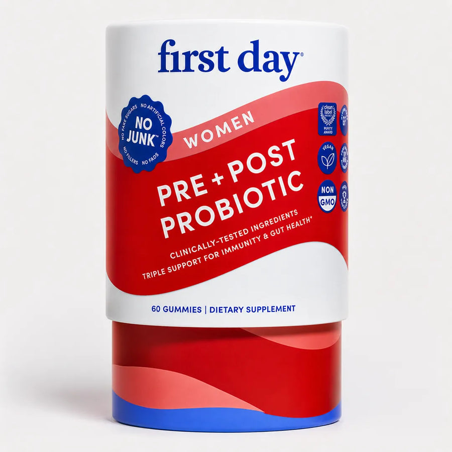 A cylindrical container of First Day Women Pre + Post Probiotic dietary supplement gummies, featuring a white, red, and blue label on a light gray background.