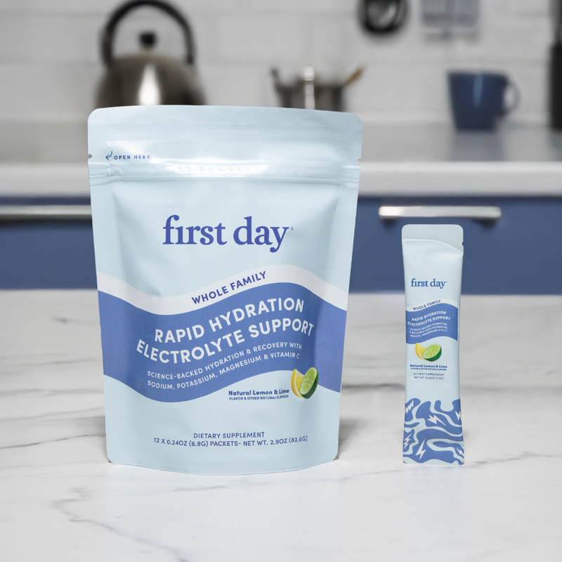 A pouch and a single stick packet of First Day Rapid Hydration Electrolyte Support with Natural Lemon & Lime flavor are displayed on a white kitchen counter with a blue and white background.