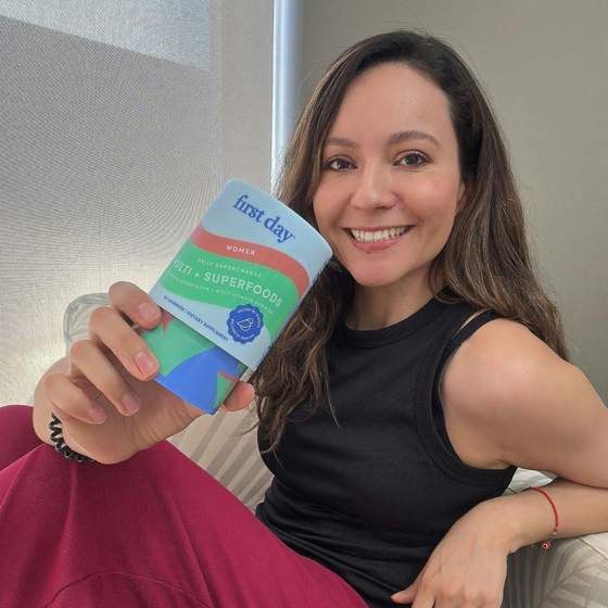 A smiling woman with long brown hair, wearing a black sleeveless top and red pants, holds up a package labeled 1st day women multivitamin superfood. She is sitting indoors near a window.