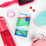 A bottle of First Day Women The Multi dietary supplement is displayed in a styled flat-lay arrangement on a white background, surrounded by fitness and lifestyle props including a pink hairbrush, a red water bottle, pink hair ties, a mint sneaker, and a pink alarm clock.