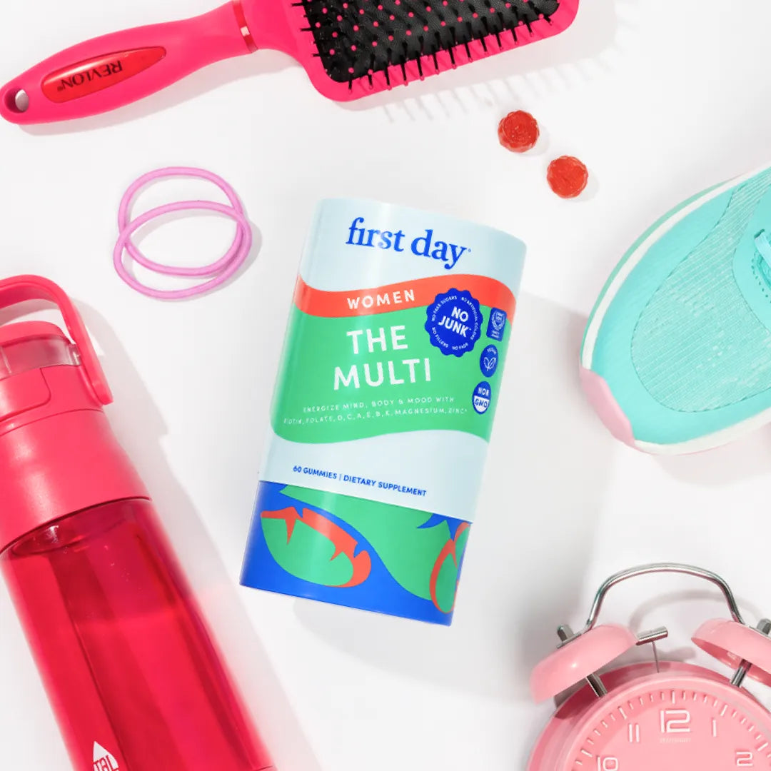 A bottle of First Day Women The Multi dietary supplement is displayed in a styled flat-lay arrangement on a white background, surrounded by fitness and lifestyle props including a pink hairbrush, a red water bottle, pink hair ties, a mint sneaker, and a pink alarm clock.