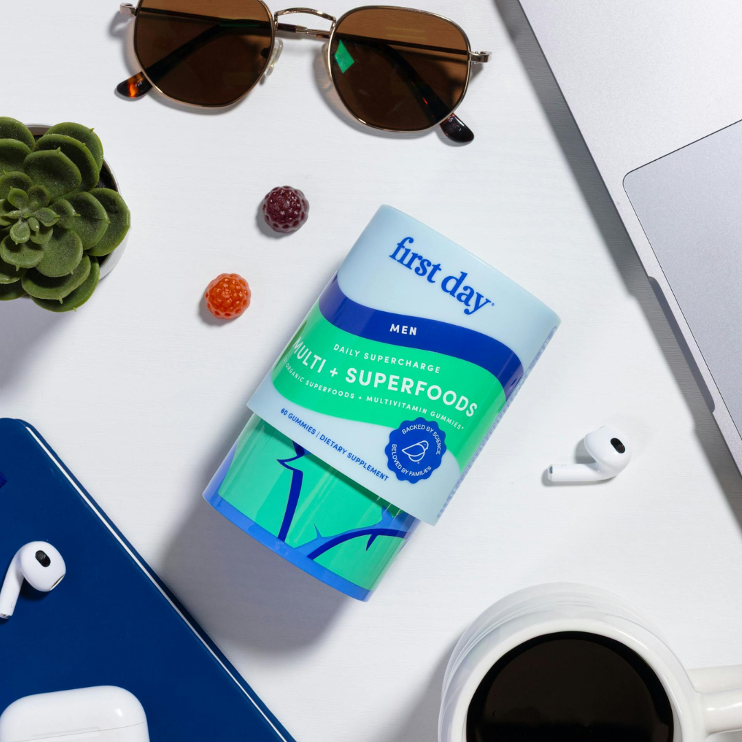 A First Day Mens Multi + Superfoods supplement bottle on a white surface, surrounded by sunglasses, earbuds, a succulent plant, gummy vitamins, a laptop, a blue notebook, and a cup of coffee.