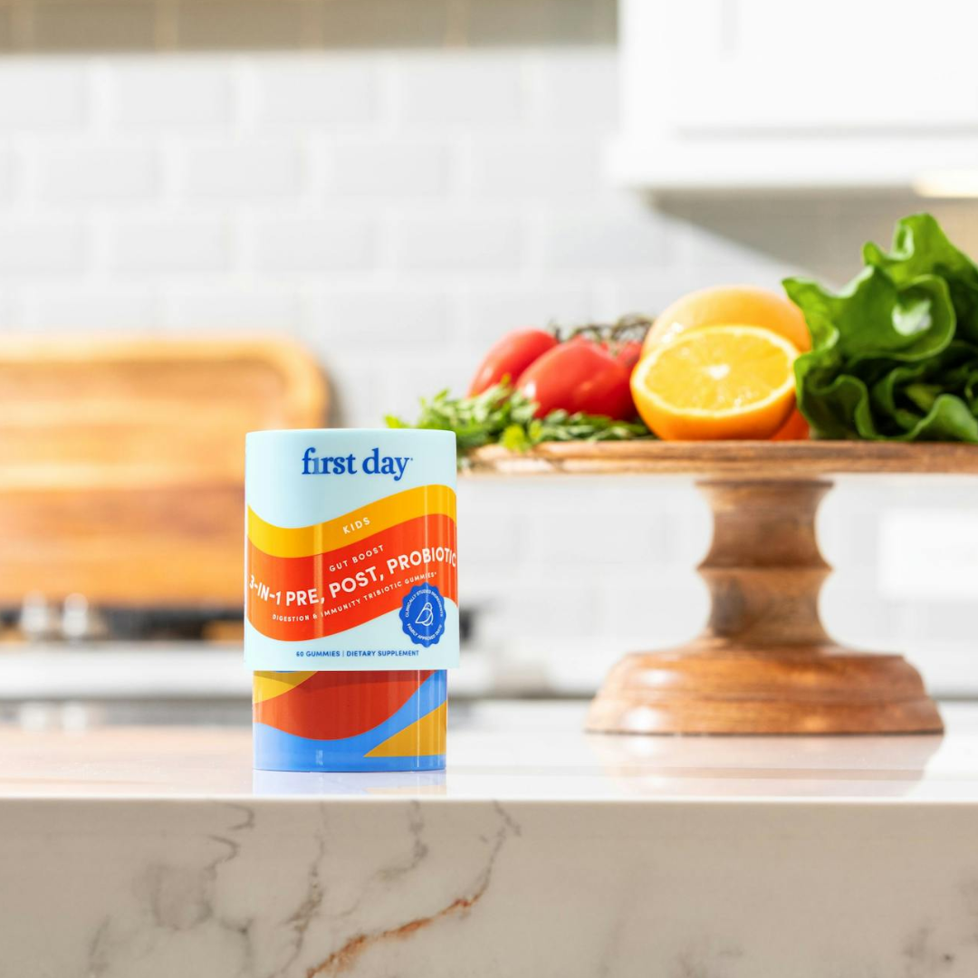 A container labeled first day kids pre, post, and probiotic sits on a marble countertop with fresh vegetables, lettuce, tomatoes, and a halved lemon on a wooden cake stand in the background.