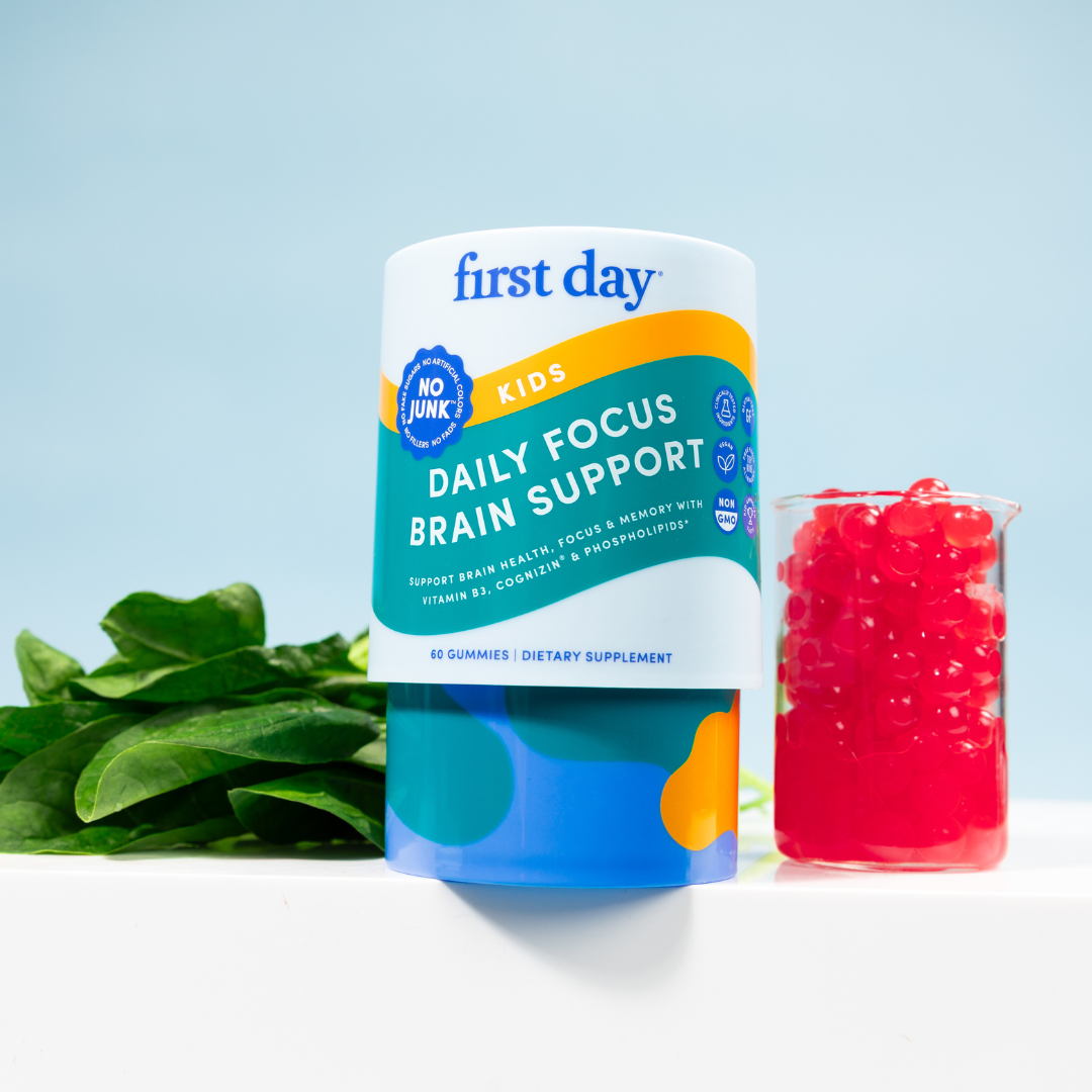 A container of First Day Kids Daily Focus Brain Support gummies stands next to fresh spinach leaves and a clear cup filled with red gummies, set against a light blue background.