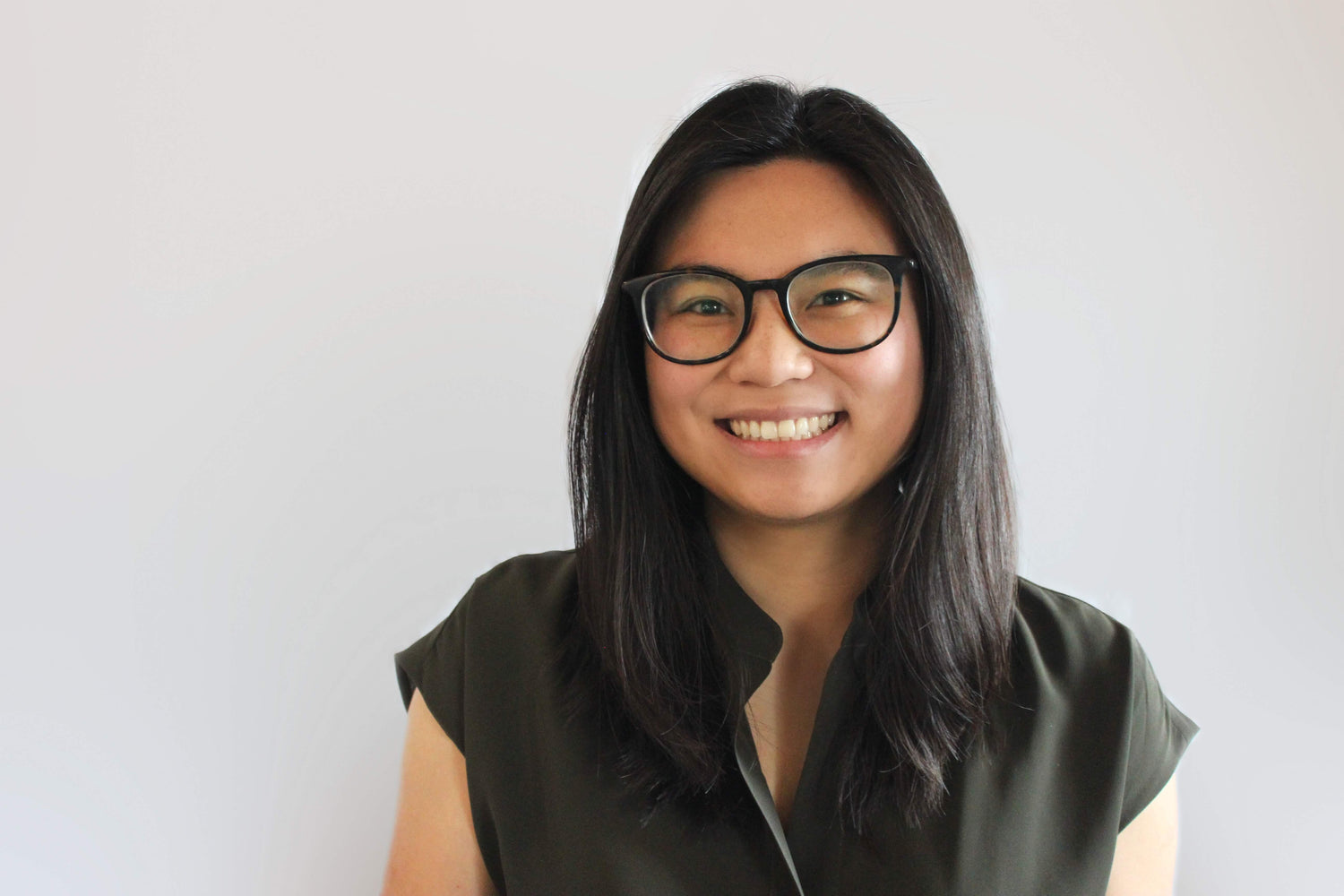 A person with long, straight dark hair and glasses smiles at the camera. They are wearing a dark short-sleeved shirt and are posed against a plain, light-colored background.