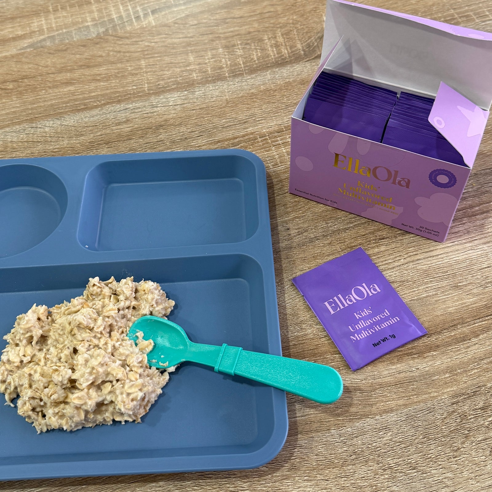 Blue divided plate with food, green spoon, and EllaOla packets on a wooden surface