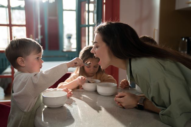 A young boy feeds a woman with a spoon as she leans forward, smiling. Two other children sit nearby, eating from white bowls at a kitchen counter in a sunlit room.