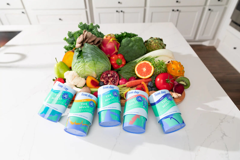 Five bottles of First Day multivitamins are displayed on a kitchen counter in front of a colorful assortment of fresh fruits and vegetables, including pomegranate, grapefruit, kiwi, and leafy greens.
