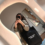 In a cozy room, someone takes a mirror selfie holding the Reflection Tote, which reads 