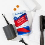 A First Day Men pre- and post-probiotic supplement bottle is arranged in a flat-lay with a toothbrush, razor, two orange gummies, a towel, and a brush on a white background.