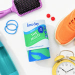 A bottle of First Day Teens The Multi daily supplement gummies is displayed in a styled flat-lay on a white background, surrounded by lifestyle props including a purple hairbrush, an orange athletic shoe, blue hair ties, a yellow alarm clock, and a teal water bottle.