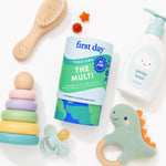 A styled flat-lay of baby and toddler products on a white background, featuring a container of First Day Toddlers 'The Multi' dietary supplement, a bottle of Gentle Wash soap, a wooden baby brush, a ring stack toy, a pacifier, and a dinosaur teether.
