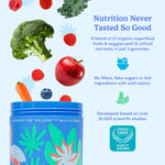 A blue tin of women's multivitamin gummies with colorful tropical leaf illustrations, shown on a light blue background with floating fruits and vegetables including broccoli, red berries, an apple, a carrot, and a blueberry. Text on the ad reads 'Nutrition Never Tasted So Good' and includes a Clean Label award badge.