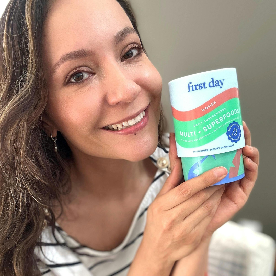 A smiling woman with long brown hair holds up a container of First Day Women’s Multivitamin, showing the label to the camera. She is indoors and wearing a striped shirt.