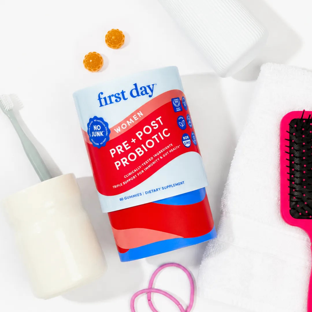 A bottle of First Day Women Pre &amp; Post Probiotic gummies is styled on a white background with bathroom accessories including a toothbrush, cotton pads, a pink hairbrush, and rubber bands.